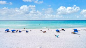 Ein Strand mit weißem Sand, klarem türkisblauem Wasser und einigen Personen. Blaue Sonnenschirme und Liegestühle sind vorhanden.