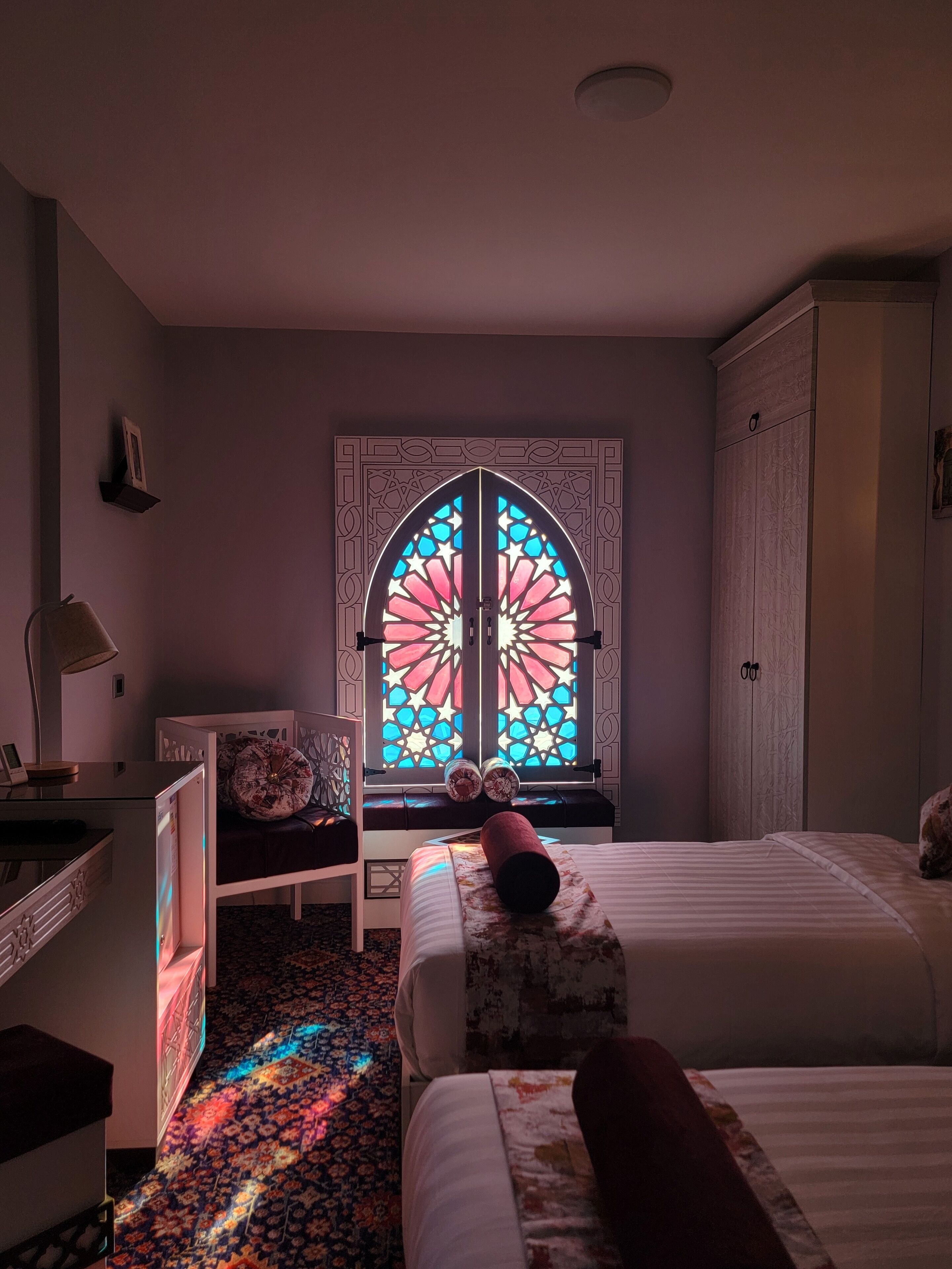 A room with a bed, a desk, a chair, and a window with a stained glass design.