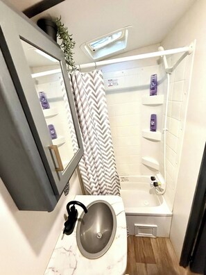 Ein kleines Badezimmer mit einem weißen Waschbecken, einem Spiegel, einer Dusche mit Vorhang in Zickzackmuster und Ablagen mit Pflegeprodukten.
