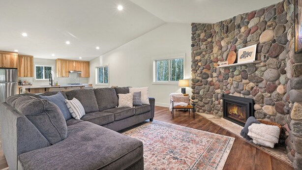 A modern living room with a stone fireplace, a gray sectional sofa, and a kitchen in the background. - Book Your Mt. Baker Après Ski Stay (Deming)