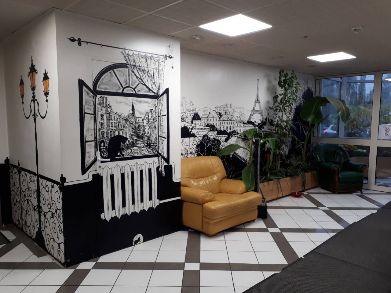 A lobby with a checkered floor, a brown leather chair, a large potted plant, and a mural depicting a cityscape.