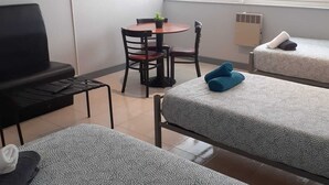 Ein Zimmer mit zwei Betten, einem kleinen Tisch mit Stühlen, einem an der Wand montierten Fernseher und einem Fenster mit Jalousien.