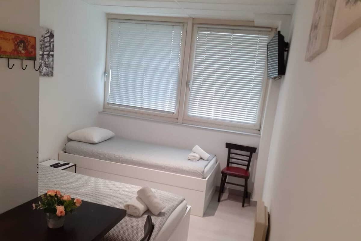 A small, compact room with a bed, a chair, a table, and a window with blinds.