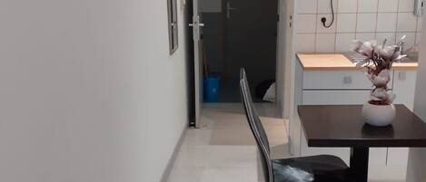 A compact hotel room with a bed, a small table with a vase, a chair, a kitchenette with a microwave, and a door leading to a bathroom.