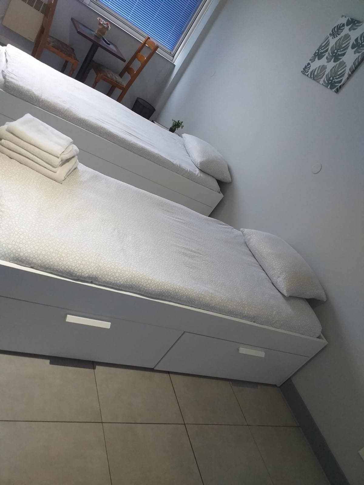 A room with two beds, a small table, and a window with blinds.