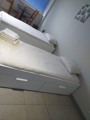 A room with two beds, a small table, and a window with blinds. - Appart Hotel (Champagne-sur-Oise)