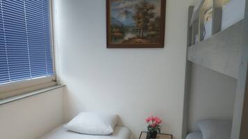 A small, clean room with a single bed, a small table with a vase, and a painting on the wall.
