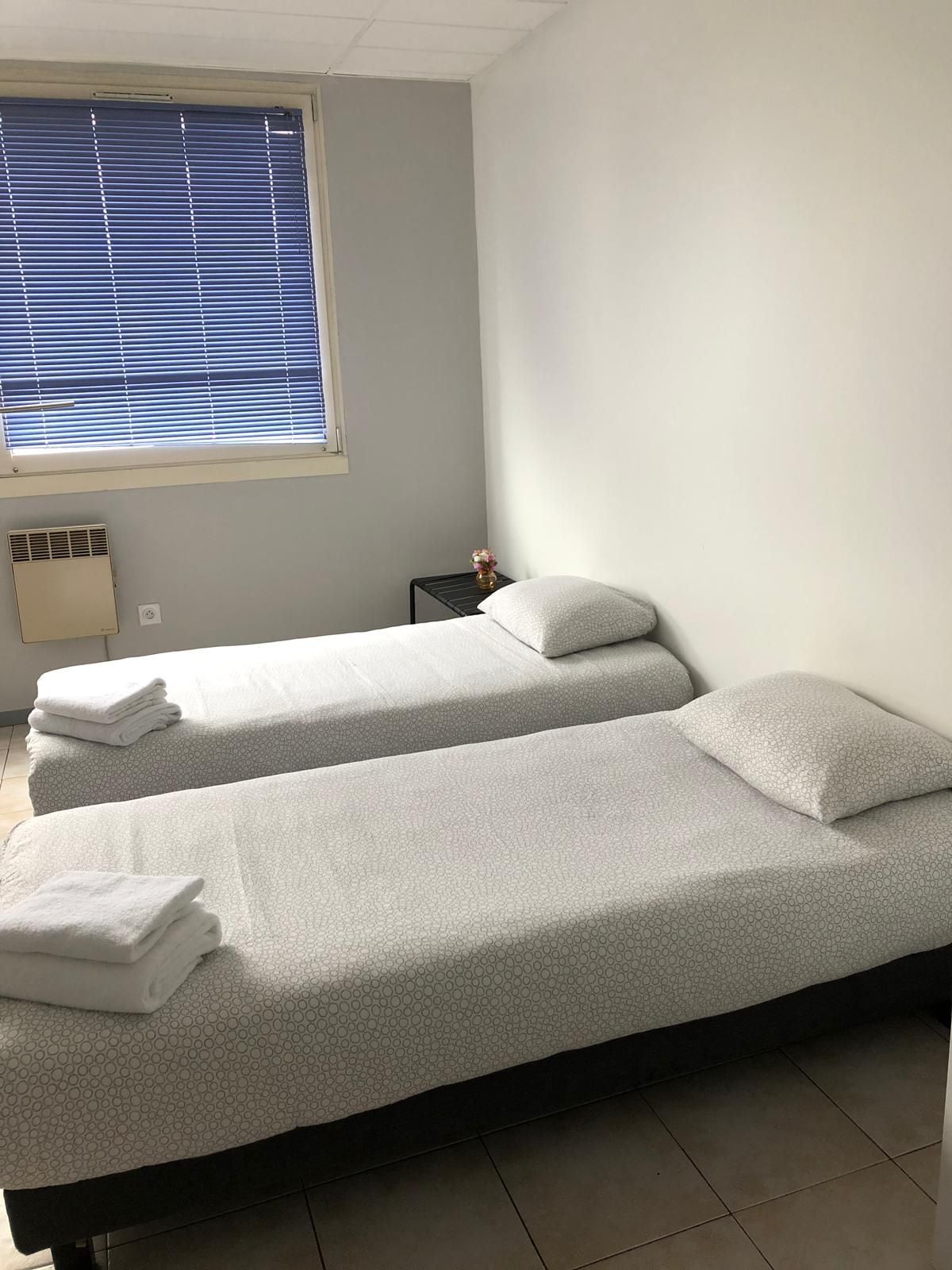 Een kamer met twee éénpersoonsbedden, een klein nachtkastje met een vaas, een raam met jaloezieën en een wandradiator.