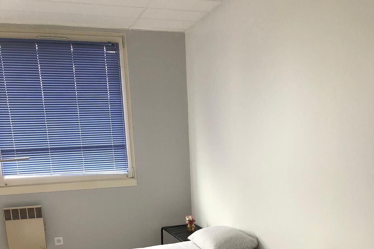 A room with two single beds, a small bedside table with a vase, a window with blinds, and a wall-mounted heater.
