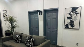 Een woonkamer met een grijze bank, twee gekleurde sierkussens, een potplant en twee donkere deuren.