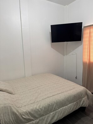 A room with a bed, a flat-screen TV mounted on the wall, and a window with curtains.