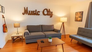 A living room with a grey sofa, a wooden coffee table, a grey armchair, a guitar, and a wall sign that reads "Music City". - The Eastside 2BR Near 5Points (Nashville)
