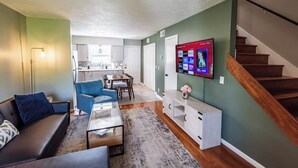 A modern living room with a sofa, coffee table, and TV. There is a staircase to the right and a kitchen in the background. - Riverside Retreat Near 5 Points (Nashville)
