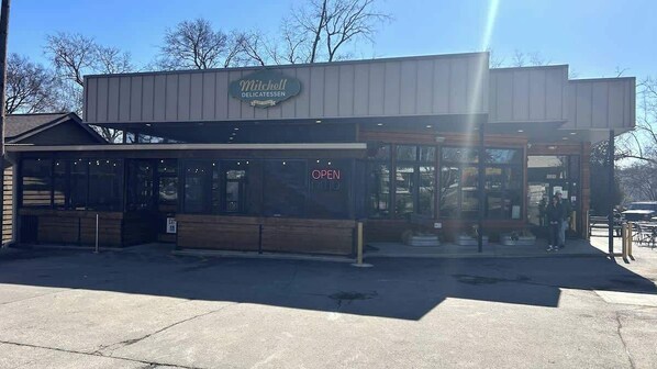 Een restaurant genaamd Mitchell Delicatessen met een 'OPEN' bord, terras en een heldere lucht.