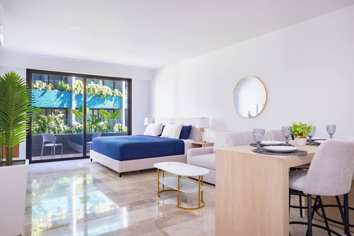 Una habitación de hotel moderna con una cama, una mesa de comedor con sillas, una mesita blanca pequeña y un ventanal con vistas a la vegetación.