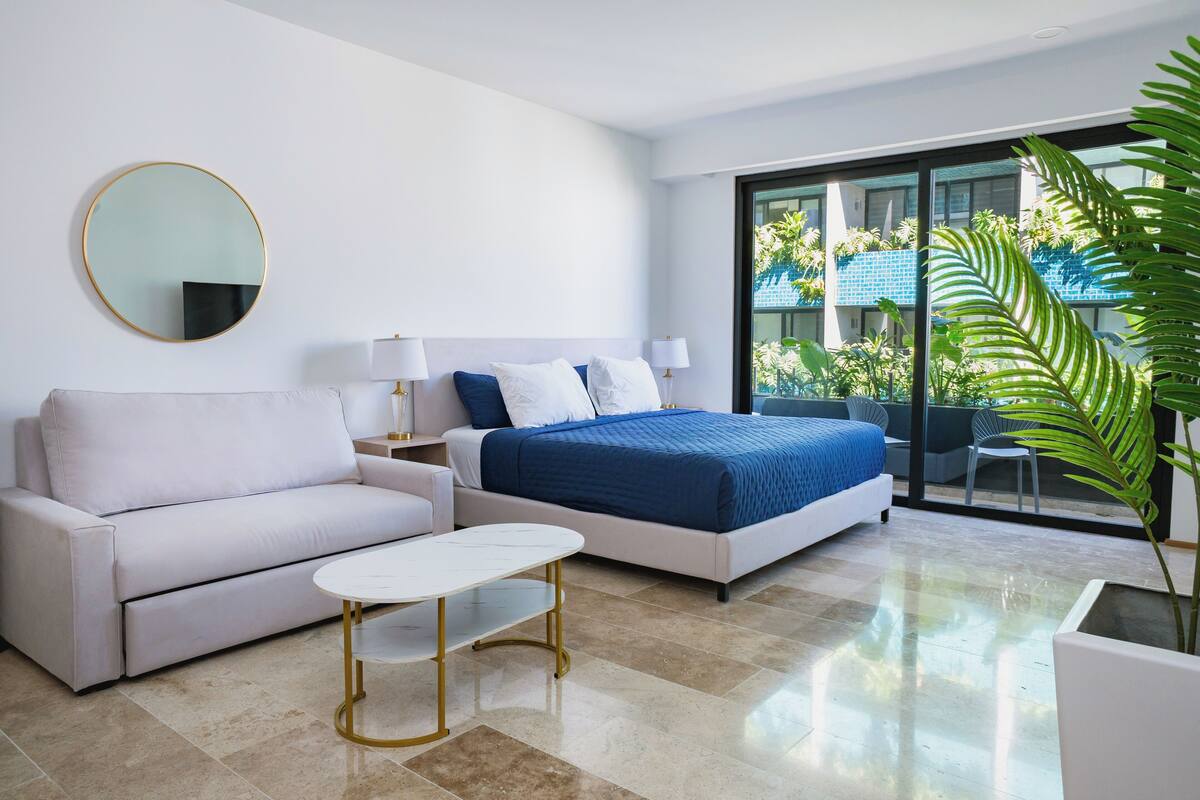 Habitación de hotel moderna con cama, sofá, una mesita y un amplio ventanal con vista a zonas verdes.
