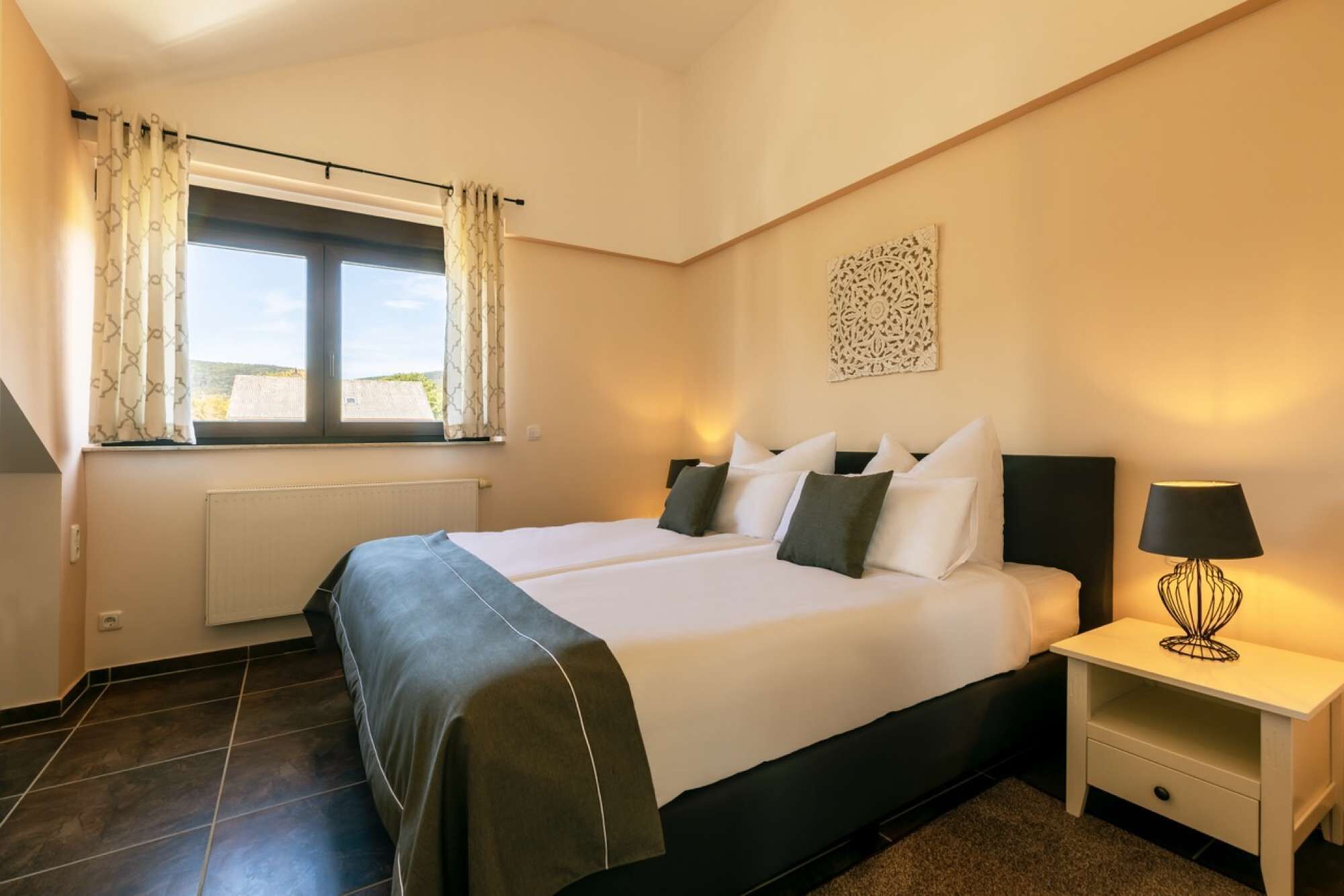 Ein Doppelbett mit schwarzem Kopfteil, ein Nachttisch mit Lampe und ein Fenster mit Vorhängen.