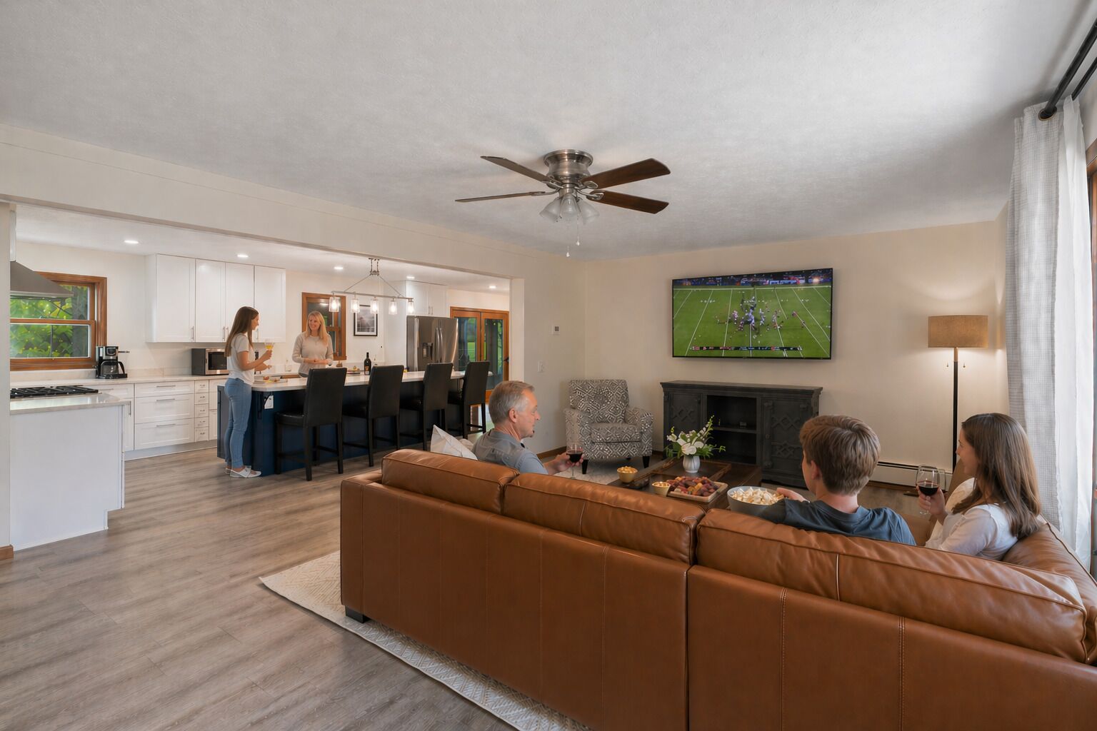 A modern living room with a brown leather sofa, a flat-screen TV mounted on the wall, and a kitchen in the background.