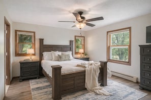 Una camera da letto con un letto di legno, comodini, una ventola a soffitto e finestre che danno su alberi.