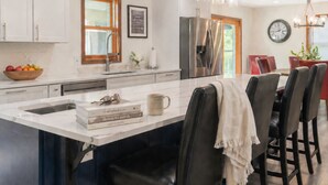 A modern kitchen with white cabinets, a large island with bar stools, a stainless steel refrigerator, and a dining area with red chairs.