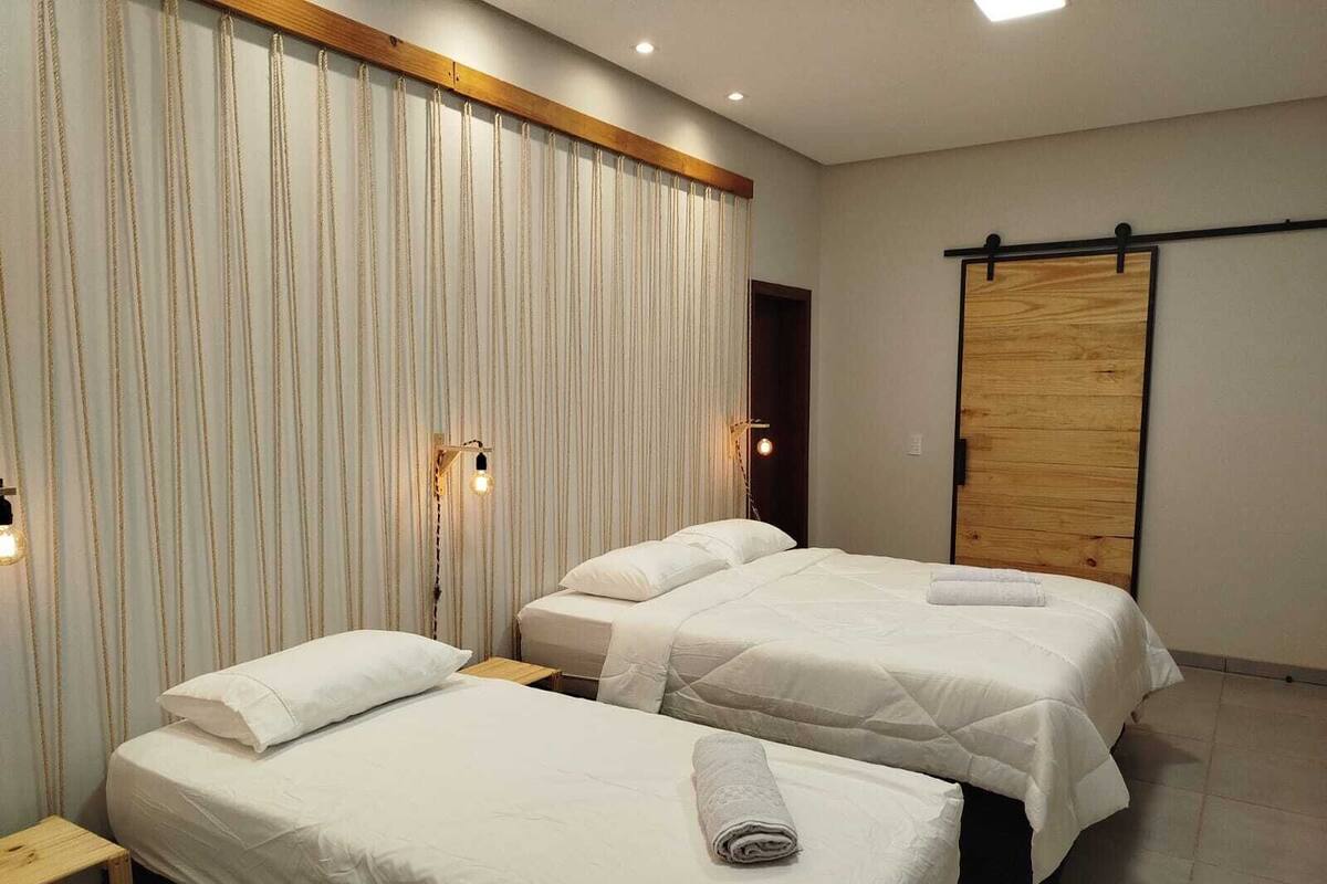 Une chambre d’hôtel avec deux lits, une tête de lit en bois, une porte coulissante et un petit tabouret en bois.