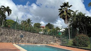 A swimming pool with a stone wall, palm trees, and a wooden bench with a towel and bottle. - Spacious apartment with pool, 5-minute walk from the beach (Le Gosier)