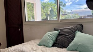 A bedroom with a bed, pillows, a window with blinds, and a door. - Spacious apartment with pool, 5-minute walk from the beach (Le Gosier)