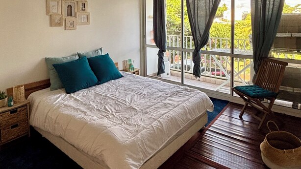 A bedroom with a large bed, a chair, a window with curtains, and a view of greenery outside. - Spacious apartment with pool, 5-minute walk from the beach (Le Gosier)