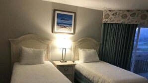 A hotel room with two beds, a nightstand with a lamp, a framed picture on the wall, and a view of the outdoors through a window with curtains.