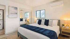 A modern hotel room with a large bed, a washing machine, a mirror, and a window with blinds.