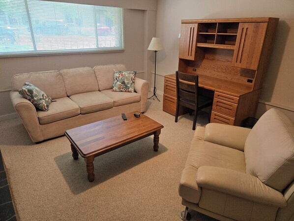Una sala de estar con un sofá color beige, una mesa de centro de madera, un escritorio con cajones y una lámpara.