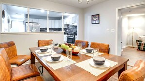 A dining area with a wooden table set for four, surrounded by leather chairs. A kitchen is visible through a large window in the background.