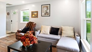 A living room with a sofa, armchair, and coffee table. There are framed pictures on the wall and a window with a view of greenery.