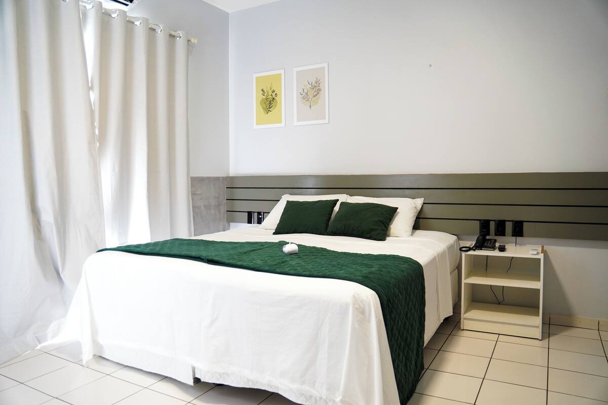 Una cama bien hecha con un plaid verde, una mesita de noche con un teléfono y dos cuadros en la pared.