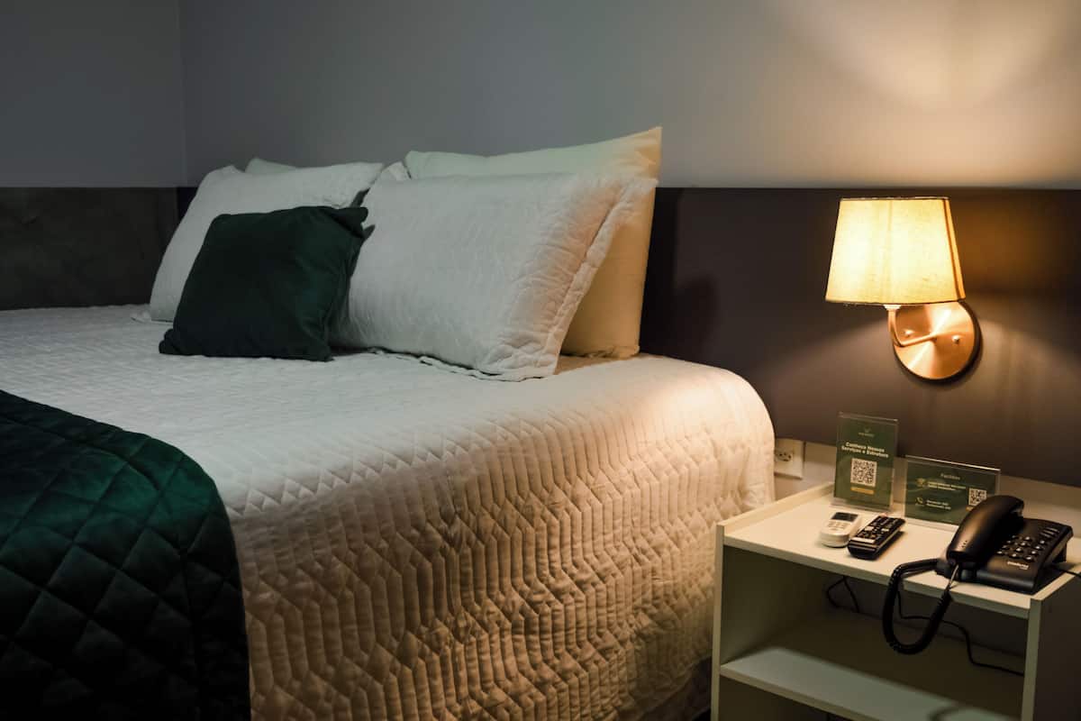 Una cama bien tendida con una colcha blanca y verde, una lámpara de noche, un teléfono y una mesita con un cartel.