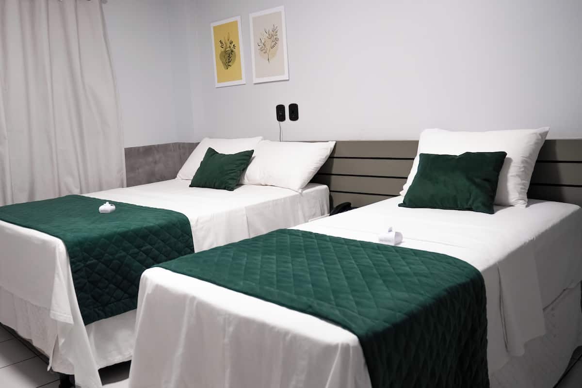 Dos camas individuales con sábanas blancas y colchas verdes, un cabecero gris y dos impresiones enmarcadas de motivos botánicos en la pared.