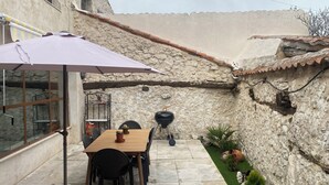 Une terrasse dallée de pierres, avec une table et des chaises en bois, un barbecue et un petit coin jardin.