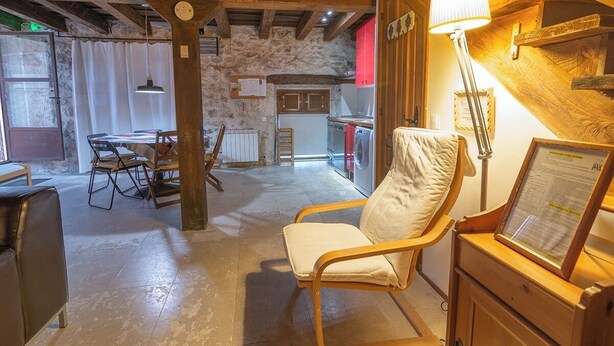 Un intérieur rustique avec des poutres apparentes, un mur en pierre, un coin repas avec une table et des chaises, et un espace détente avec une lampe et un tableau présentant le menu.