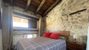 Une chambre avec un mur en pierre, un plafond en bois, un lit recouvert d’une couverture à carreaux, une table de chevet avec une lampe et une fenêtre avec des rideaux.