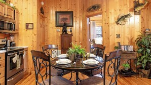 A wooden interior with a dining table set for four, surrounded by chairs. There is a kitchen area with a microwave and stove, and a door with a bear and fish decoration.