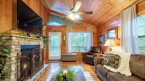 A cozy cabin interior with a fireplace, a leather couch, a television, and a ceiling fan.