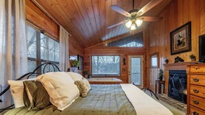 A wooden cabin bedroom with a bed, a fireplace, a ceiling fan, and a window with blinds.