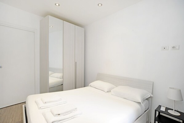 A modern hotel room with a large bed, white linens, a wardrobe, and a bedside table with a lamp.