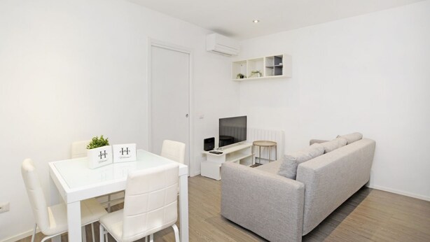 A modern, minimalist living room with a white sofa, a dining table with chairs, a flat-screen TV, and a small side table. - Ticinese 28 Apartment (MILANO)