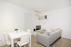 A modern, minimalist living room with a white sofa, a dining table with chairs, a flat-screen TV, and a small side table.