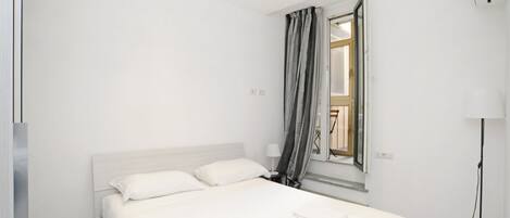Ein modernes Hotelzimmer mit einem großen Bett, einem Fenster mit Vorhängen, einem Nachttisch und einer Stehlampe.