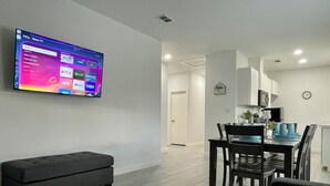 Een moderne woonkamer met een flatscreen-tv aan de wand, waarop streamingdiensten worden weergegeven. Er staat een donkergrijze bank en een eethoek met een tafel en stoelen.