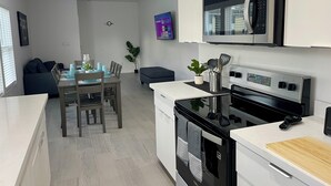 Een moderne keuken met een magnetron, oven en een houten snijplank. Er is een eethoek met een tafel en stoelen.