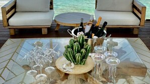 Una terraza con dos sofás orientados a una vista de aguas turquesas y una isla distante. Una mesa en primer plano tiene tapa de vidrio y base de madera, un centro de mesa con un cactus y varias botellas y vasos.