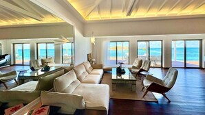 Un salón espacioso con un sofá grande, sillones y una mesa de centro. La habitación tiene ventanas amplias con vistas al océano.